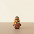 A small owl figurine wearing a knitted orange and dark blue beanie with a pom-pom and a matching red knit scarf, holding a yellow flower, standing on a wooden table.