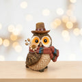 A decorative owl figurine wearing a brown hat and a red, blue, and brown knit scarf, holding small dried flowers,